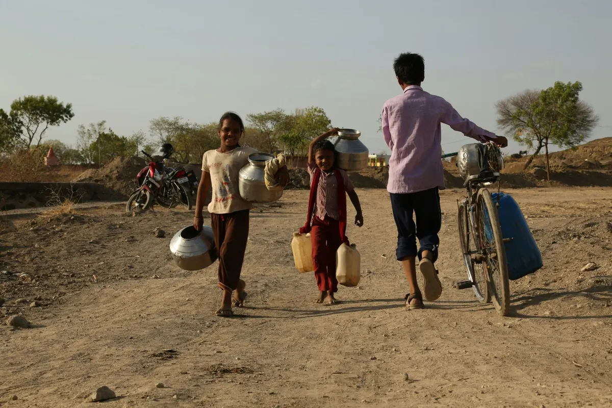 Three children carry water pots and containers along a dry dirt track